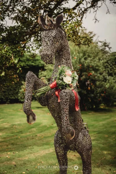 Metallic horse sculpture with floral bouquet in garden.