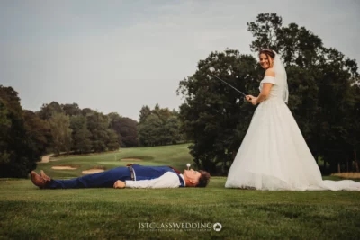 Bride posing with golf club, groom lying on grass.