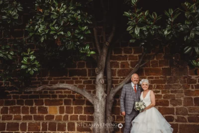 Bride and groom smiling by brick wall with tree.
