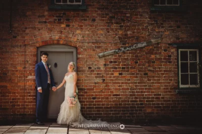 Bride and groom holding hands against rustic brick building.