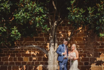 Wedding couple posing by brick wall under tree.