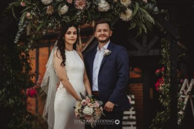 Wedding couple posing under floral archway.