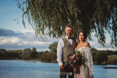 Couple posing by lake with bouquet, wedding photography.