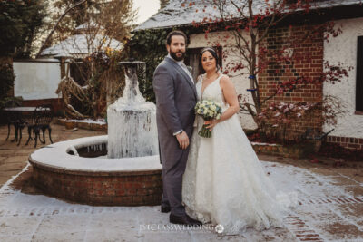 Couple posing in winter wedding attire by frozen fountain.