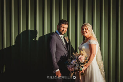Bride and groom smiling in sunlight by green wall.