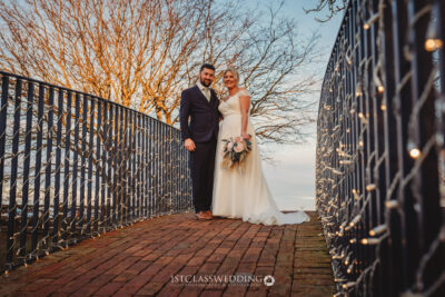 Bride and groom on bridge at sunset with fairy lights.