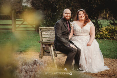 Couple in wedding attire sitting on bench outdoors.