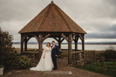 Bride and groom under wooden gazebo with coastal view.