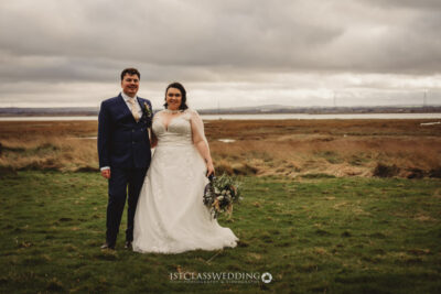 Bride and groom posing in field, overcast weather.