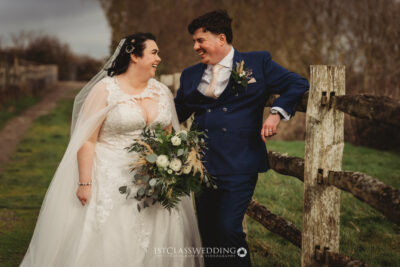 Bride and groom smiling outdoors by wooden fence.