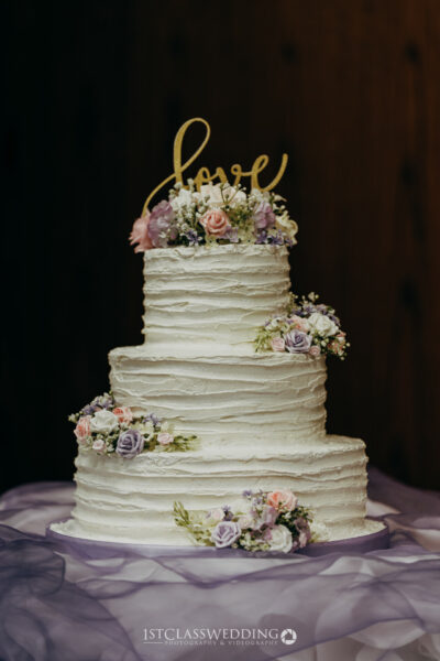 Three-tiered wedding cake with "Love" topper and flowers.
