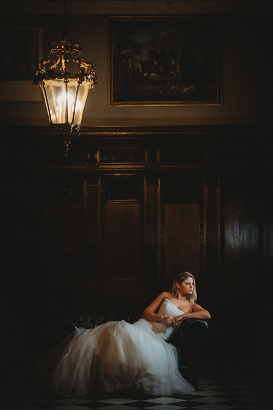 Bride in gown sitting pensively in dimly lit vintage room.