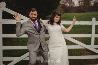 Bride and groom posing playfully by white gate outdoors.