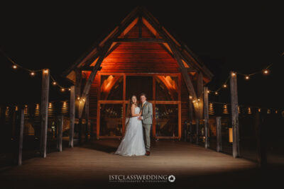 Couple posing at night, illuminated wooden venue entrance.