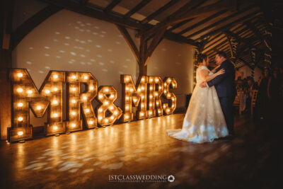 Newlyweds dancing by illuminated 'MR & MRS' sign.